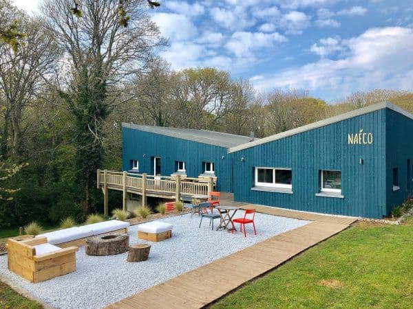 Vue exterieure sur les batiments en bois peint en bleu et la grande terrasse. celle ci à un sol en gravier blanc, est entouré de dalles en bois. Vous pourrez vou sinstaller dans de grands canapés en bois