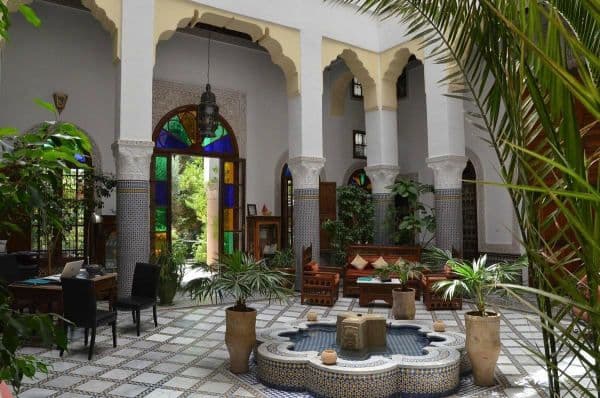 Patio traditionnelle d'un riad avec au centre une fontaine et autour des murs blancs avec un soubassement en mosaique comme sur le sol. Le bureau d'accueil y est installée dans l'angle gauche