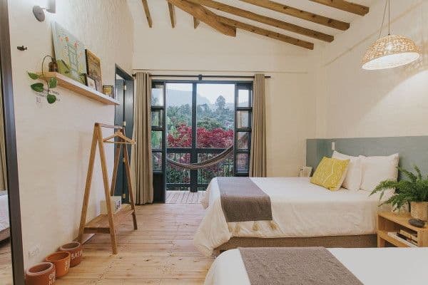 Photo prise dans une chambre à la décoration moderne et épurée. Il y a 2 grands lits et la baie vitrée ouverte donne sur un balcon où on aperçoit un hamac.