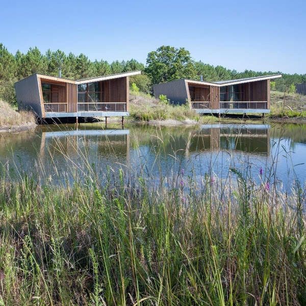 Photo de deux écolodges construit en bois et installé en partie sur des pilotis sur un étang. Ils sont de forme géométriques modernes et belles terrasses