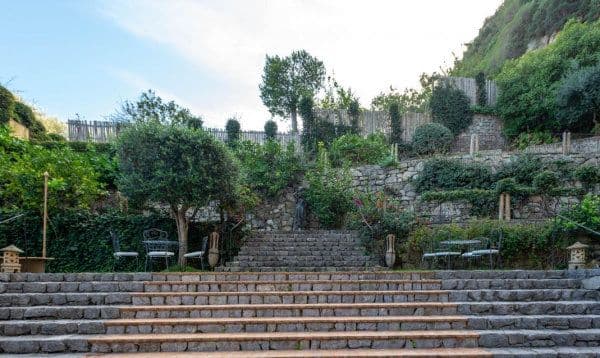 Naples - Conca Park. Jardin de l'hotel arboré en terrasse avec des marches en pierre. des tbales et chaises sont installés a quelques endroits
