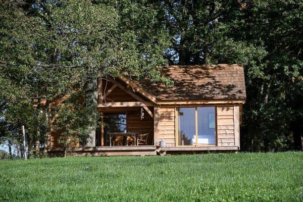 Photo d'un des lodge en bois avec une grande baie vitrée. A sa gauche un grand chene et devant une belle pelouse verte