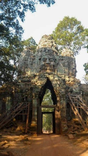 Porte ouest pour entrer sur le site Angkor. Un chemin en terre battue et entouré d'arbres passe en dessous. La porte est soutenue par des échafaudage car en cours de rénovation et sur le dessus on voit distinctement les visages