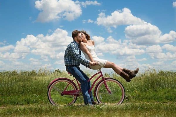 Couple sur un vélo rouge le long d'un champ fleuri. L'homme pédale et la femme est assise sur le guidon en l'embrassant