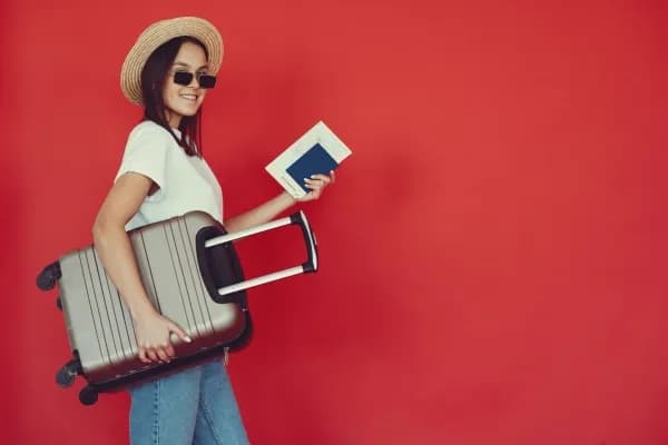 Femme avec un tee short blanc, un chapeau de paille et des lunettes de soleil portant dans une main son passeport et sous l'autre bras sa valise cabine