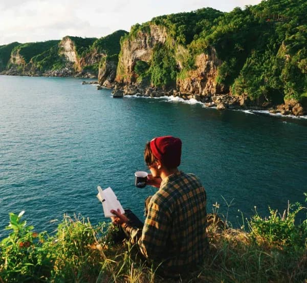 Un homme assis au bord d'une falaise fase à la mer un livre dans une main et une tasse de café dans l'autre