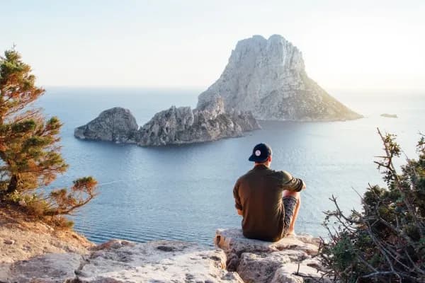 Homme casquette à l'envers assis sur un promontoire rochaux admirant la mer et une ile rocheuse face à lui
